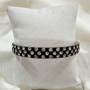 May include: A black bracelet adorned with two rows of clear rhinestones. The bracelet is presented on a white, textured surface. This piece of jewellery is a fashionable accessory.