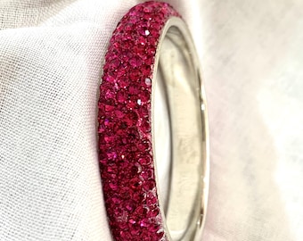 May include: A circular bangle bracelet featuring a thick band encrusted with numerous small, vibrant fuchsia pink crystals. The interior of the bracelet is a smooth, polished silver-toned metal, providing a contrast to the textured, sparkling exterior surface.