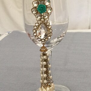 May include: An ornate wine glass charm featuring a large, clear teardrop-shaped jewel, surrounded by smaller clear stones. A teal rose is centered in the design. The charm has a gold-colored metal frame and a dangling strand of pearls.