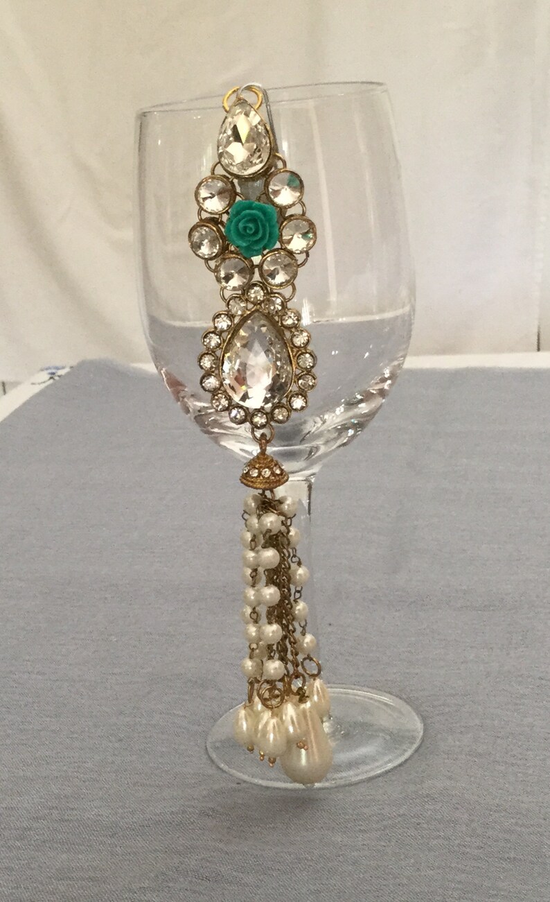 May include: An ornate wine glass charm featuring a large, clear teardrop-shaped jewel, surrounded by smaller clear stones. A teal rose is centered in the design. The charm has a gold-colored metal frame and a dangling strand of pearls.