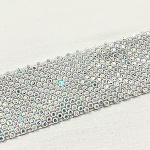 Puede incluir: Una cinta blanca con ribetes de brillantes iridiscentes. Los brillantes están dispuestos en un patrón de cuadrícula.