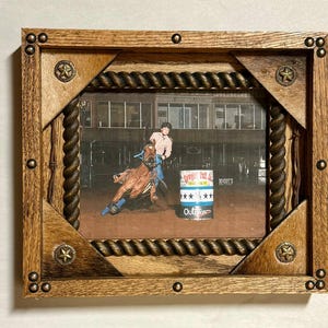 May include: A rectangular wooden picture frame with a rope-textured inner border, metal star accents at the corners, and decorative studs. The frame contains a photograph of a person riding a horse around a barrel labeled Cowgirl Tuff Co. and Outlaw.