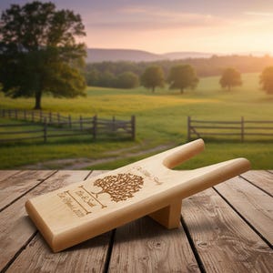 May include: A light-colored wooden boot jack featuring a laser-engraved design of a tree, the text "The Miller Family", "Erin & Sarah", and "Est. March 8, 2020". The boot jack has a U-shaped cutout at one end and a support block underneath.
