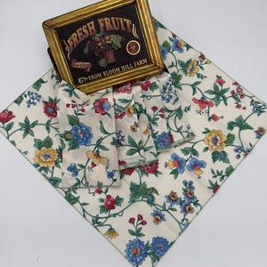 Può includere: Tovaglia e tovaglioli con motivo floreale. Lo sfondo è color crema con fiori rossi, blu e gialli e foglie verdi. Un quadro con la scritta "Fresh Fruit" è presente nell'immagine.