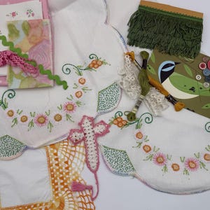 May include: A collection of craft supplies including white embroidered linens with floral patterns, a yellow crocheted square, green fringe trim, pink rickrack, embroidery floss, a cross-shaped lace applique, and a card with buttons.