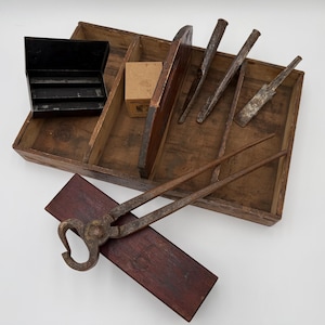 Puede incluir: Una bandeja de herramientas de madera con varias herramientas de metal, incluyendo cinceles, una tenaza y una pequeña caja negra. La bandeja también contiene una pequeña caja de cartón. Las herramientas están dispuestas sobre una superficie blanca.