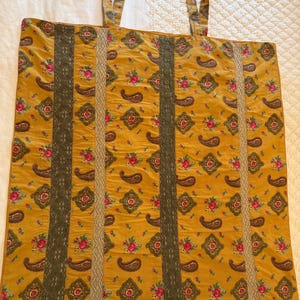 May include: A mustard yellow fabric panel featuring a repeating pattern of pink roses, green leaves, and dark paisley shapes. The panel is accented with vertical strips of dark olive green lace and light beige textured ribbon trim.