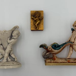 May include: Three decorative figurines. One is a cream-colored vase held by two cherubs. Another is a gold-colored rectangular relief of a cherub playing a harp. The third is a cherub pushing a decorative wave.