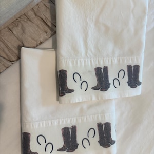 May include: Two white pillowcases with a border featuring brown cowboy boots and black horseshoes. The pillowcases are folded and arranged on a white surface, showcasing a rustic, country theme.