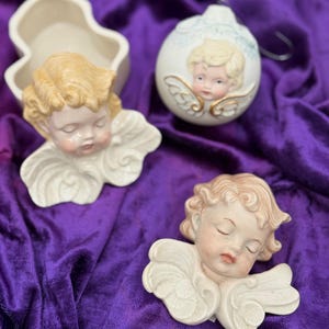 Könnte beinhalten: Drei Keramik-Puttenfiguren mit geschlossenen Augen und detaillierten Flügeln. Eines ist ein Weihnachtsschmuck, ein anderes ist ein Wandbehang, und das dritte ist ein kleiner, offener Behälter. Die Putten haben blondes Haar und sind vor einem violetten Samthintergrund platziert.