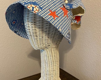 Puede incluir: Un sombrero para el sol a cuadros azules y blancos con diseños náuticos, que incluyen estrellas rojas, salvavidas y anclas. El sombrero se exhibe en una cabeza de maniquí de mimbre blanco.