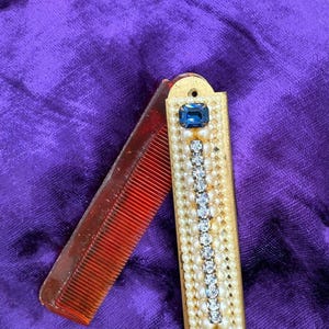 May include: A vintage folding comb. The red plastic comb is open, revealing its teeth. The gold-coloured case is decorated with faux pearls, clear rhinestones, and two blue rectangular stones. The comb rests against a purple background.