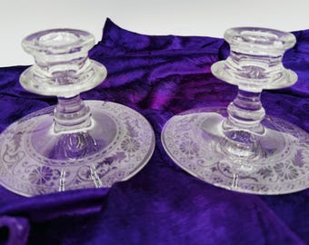 May include: Two clear glass candle holders with a decorative floral pattern etched on the base. The candle holders are displayed on a deep purple velvet fabric. The candle holders have a classic design.