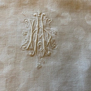 Op de afbeelding: Wit linnen stof met een delicaat geborduurd monogram in het midden. De stof heeft een subtiel stippenpatroon.