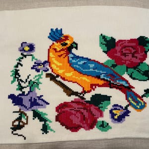May include: White fabric with a cross-stitched design featuring a colorful parrot perched on a branch, surrounded by roses and other flowers. The parrot has orange, blue, and yellow feathers. The flowers are red, purple, and blue.
