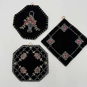 May include: Three black, embroidered pot holders with floral designs. One features a basket of flowers, while the others have repeating floral patterns. Each has a wooden ring for hanging and white stitching.