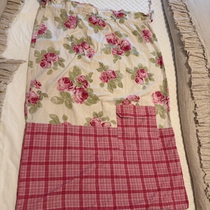 May include: A floral apron with a cream-colored background and pink roses. The bottom half features a red and white plaid pattern, with a small pocket. The apron is designed for kitchen use.
