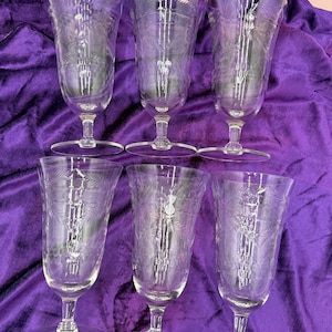 Könnte beinhalten: Sechs klare Wassergläser mit Stiel und geätzten floralen Designs. Die Gläser haben einen ausgestellten oberen Rand, einen schlanken Stiel und einen runden Fuß. Die Gläser sind auf einer violetten Samtoberfläche angeordnet.