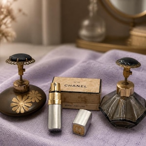 May include: A collection of vintage perfume bottles and a spray perfume bottle on a lavender surface. The bottles feature elaborate designs, including gold detailing and floral motifs. A small box of Chanel perfume is also visible.
