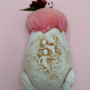 Könnte beinhalten: Ein Vintage-Nadelkissen mit einem rosafarbenen Samtbezug und einer weißen Porzellanbasis. Die Basis zeigt ein Relief von Cherubinen und floralen Designs. Eine kleine rote Blume und Nadeln sind oben befestigt.