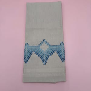 May include: A folded, light-green towel with a detailed blue embroidered design. The design features a central geometric shape with symmetrical leaf-like patterns on either side. The towel has a textured appearance and a decorative border.