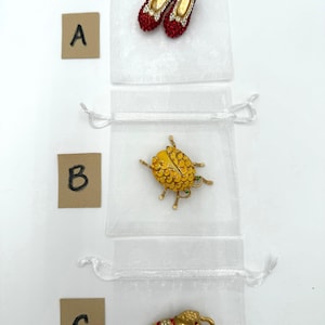 Peut inclure: Trois petits sacs blancs à cordon contiennent chacun un bijou différent. Le sac supérieur contient une paire de chaussures rouges ornées de bijoux. Le sac du milieu contient une broche en forme de scarabée jaune. Le sac inférieur contient une broche éléphant dorée.