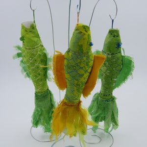 Puede incluir: Tres adornos de pescado de tela hechos a mano con tela verde y amarilla, colgados de un soporte de alambre. Los peces tienen ojos de cuentas y están decorados con una variedad de texturas.