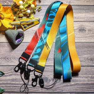 Può includere: Tre lacci colorati con diversi motivi. I lacci sono realizzati in tessuto intrecciato e hanno un moschettone in metallo nero all'estremità. I lacci sono su una superficie di legno.