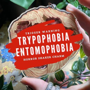 Op de afbeelding: Een heldere, op horror geïnspireerde shaker-bedel met een bruin en rood ontwerp. De bedel heeft de woorden "TRIGGER WARNING TRYPOPHOBIA ENTOMOPHOBIA HORROR SHAKER CHARM" in witte tekst op een rode achtergrond.
