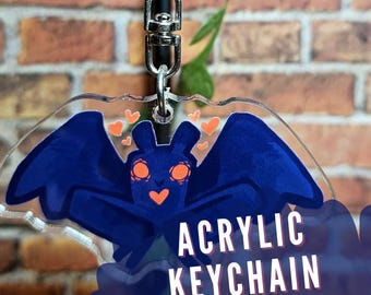 May include: A blue and orange cartoon bat with hearts around it is printed on a clear acrylic keychain. The keychain is 2 inches tall and has a flat finish. "Acrylic Keychain" and "Flat Finish | 2 Inches Tall" are printed on the bottom of the keychain.