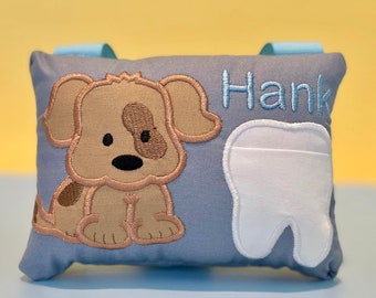May include: A gray tooth pillow with a brown and white embroidered puppy and the name "Hank" embroidered in blue. The pillow has a white tooth-shaped pocket.