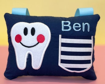 May include: A navy blue pillow with a white tooth with a smiling face and red cheeks, a white pocket with navy blue stripes, and the name "Ben" embroidered on it.
