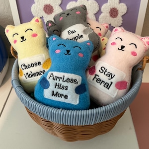 May include: A woven basket holds several plush cat toys in various colors, including yellow, blue, gray, and pink. Each cat has a smiling face and a small sign with text like "Choose Violence" and "PurrLess, Hiss More". A floral design is in the background.