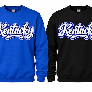 May include: Two crewneck sweatshirts, one in royal blue and one in black, each featuring the word Kentucky printed across the chest in a white script font with a blue outline. Both garments have long sleeves and ribbed cuffs and necklines.