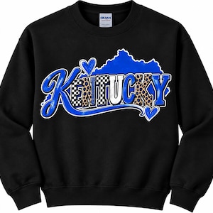 May include: A black crewneck sweatshirt featuring a graphic of the state of Kentucky in blue. The word KENTUCKY is written in a stylised font with blue, black and white chequered, and leopard print patterns, accented by two blue hearts.