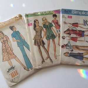 May include: Three vintage Simplicity sewing pattern packets. The packets feature illustrations of women in various outfits, including dresses, pantsuits, and skirts. The packets are labeled with the Simplicity logo and pattern numbers.