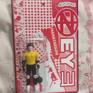 May include: A packaged action figure with a yellow shirt, black pants, and a black ball and chain. The packaging is red and white with the word "EYE" and "HANATARASHI" printed on it. The figure is in a clear plastic case.