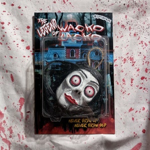 May include: A plastic package with a white, black, and red background. The package features a hanging doll head with long black hair, white skin, and red lips. The text on the package reads "The Legend of Wacko Jacko" and "Never Grow Up... Never Grow Old".