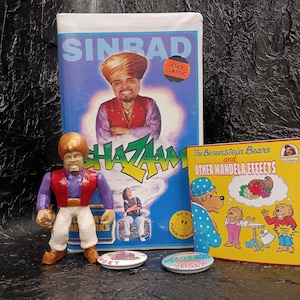 May include: A Sinbad Shazaam VHS tape, a custom action figure of a genie in a red vest and white pants, a yellow book titled The Berenstein Bears and Other Mandela Effects, and two small round pinback buttons with text on a black textured surface.