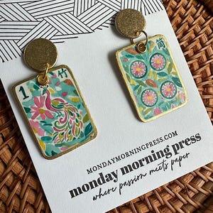 May include: A pair of rectangular drop earrings featuring gold-toned circular studs and dangling tiles with colorful illustrations. One tile displays a peacock and the number 1, while the other shows floral patterns and Chinese characters.