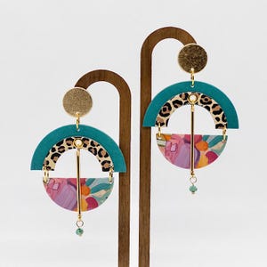 May include: A pair of statement earrings with a geometric design. The earrings feature a teal, leopard print, and floral pattern. The earrings are suspended from gold-toned hoops.