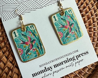 May include: A pair of rectangular drop earrings featuring a colourful illustration of a bird, bamboo stalks, and the number 1 on a light blue background. The earrings have gold-toned metal hooks and are displayed on a white card with black text.