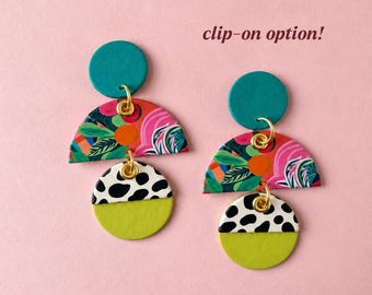 May include: Dangle earrings featuring a teal circle top, a semi-circle with a colorful floral pattern, and a bottom circle with a black and white spotted design and a solid lime green base. The text clip-on option is visible.