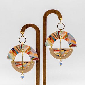 May include: A pair of colorful, geometric earrings with a semi-circle shape. The earrings are made of wood and have a gold-tone finish. Each earring has a small blue crystal dangling from the bottom.