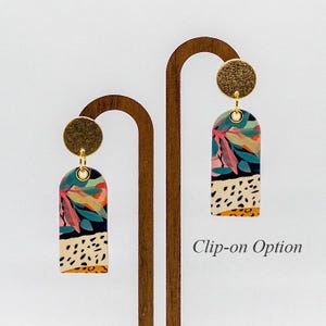 May include: A pair of clip-on earrings with a gold-tone setting and a rectangular dangle featuring a colorful floral and animal print design. "Clip-on Option" is written in the image.