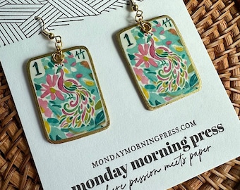May include: A pair of rectangular dangle earrings featuring a colourful peacock illustration on a light blue background with pink flowers, the number 1, and a Chinese character. The earrings have gold-toned metal hooks and edges.