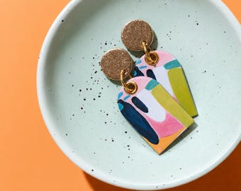May include: A pair of dangle earrings featuring gold-toned circular studs connected to arch-shaped pendants. The pendants display a colorful abstract pattern with shades of pink, lime green, dark blue, and white, attached by gold-toned jump rings.