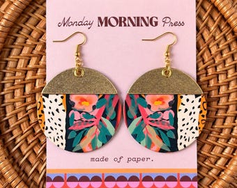 May include: A pair of round dangle earrings featuring a gold-toned top section and a colorful floral and animal print pattern on the lower portion. The earrings are displayed on a light pink card labeled Monday MORNING Press made of paper.