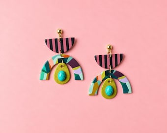 May include: A pair of dangle earrings featuring a gold-tone stud attached to a black and pink striped semi-circle. Below hangs a multi-coloured geometric arch in shades of green, purple, and orange, centred with a small, oval, turquoise-coloured bead.