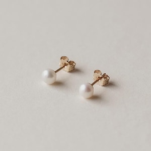 May include: A pair of gold stud earrings, each with a small, round, white pearl. The earrings are set on gold posts with butterfly clasps. The background is a soft, neutral colour.